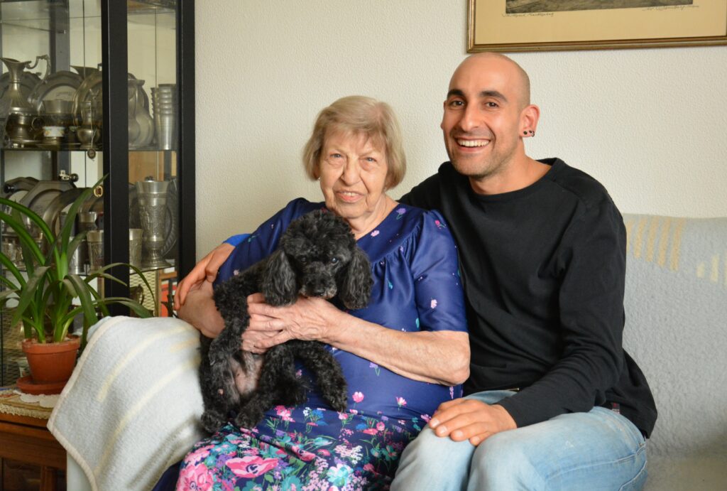 Ältere Frau mit Hund auf den Schoß sitzt neben einem ehrenamtlichen Unterstützer auf einem Sofa
